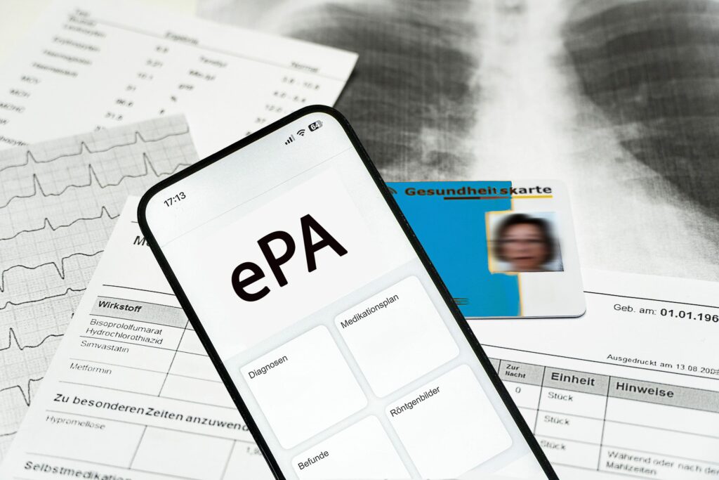 Smartphone im Vordergrund mit ePA Elektronische Patientenakte. Im Hintergrund Gesundheitskarte und Papiere.